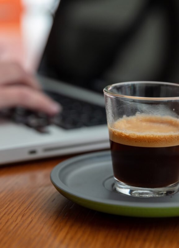 Fresh espresso coffee on a wooden office desk close up view. Woman work with a computer laptop. Fresh espresso coffee on a wooden desk close up view. Blur female hands on a computer laptop keyboard, Morning at the office